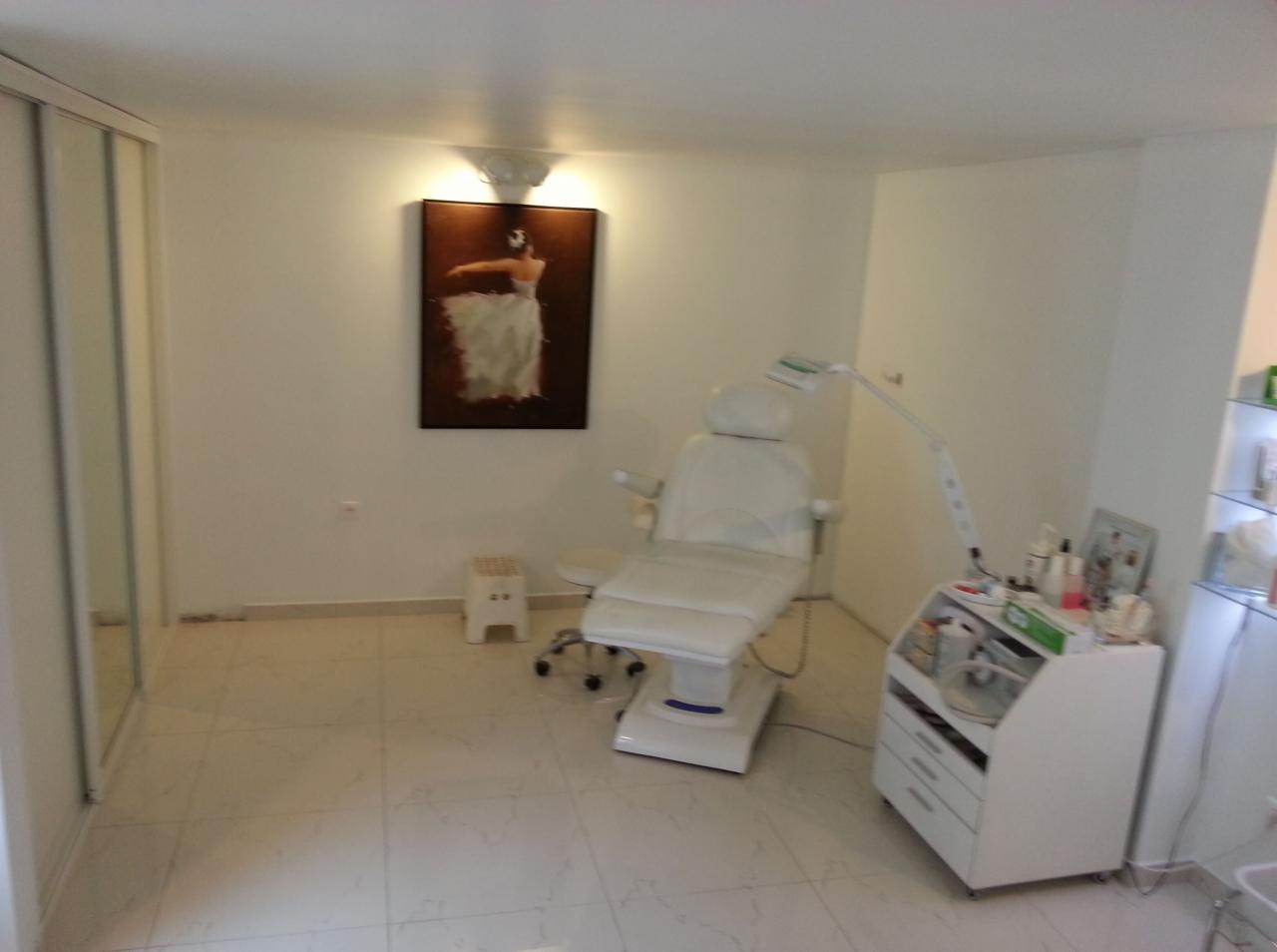 Biały, regulowany fotel kosmetyczny z oświetleniem, mały taboret i szafka z kosmetykami w jasnym gabinecie z obrazem tancerki na ścianie.