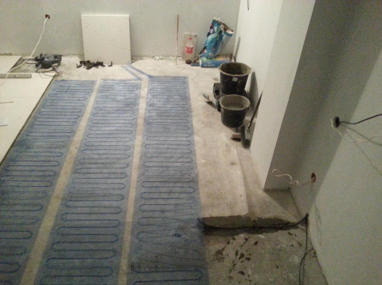 Montaż elektrycznego ogrzewania podłogowego w pomieszczeniu, widoczne maty grzewcze z niebieskim przewodem na surowym podłożu, obok wiadro i narzędzia.