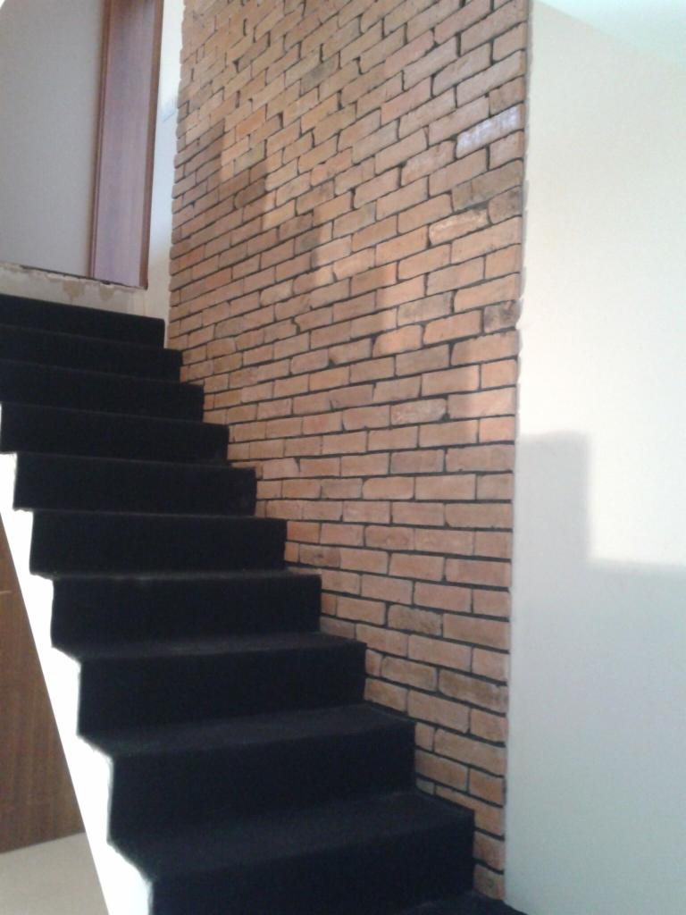 Czarne schody dywanowe przylegają do ściany z cegły dekoracyjnej w odcieniach czerwieni i brązu. Widoczny fragment drzwi w tle.