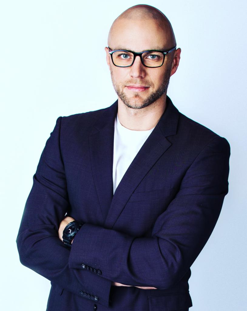 Portret mężczyzny w okularach, ubrany w granatowy garnitur i białą koszulkę, z założonymi rękami, na jasnym tle.