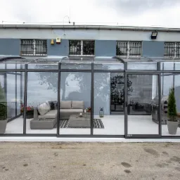 Zadaszony taras z aluminiową konstrukcją i przeszklonymi ścianami, wyposażony w szare meble wypoczynkowe, stolik kawowy, rośliny w donicach, grill i dekoracje, widok z zewnątrz.