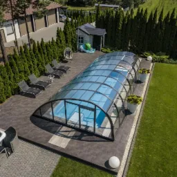 Zadaszony basen z przesuwną konstrukcją na posesji z zielonym trawnikiem, widok z góry, obok jacuzzi, leżaki i altanka.
