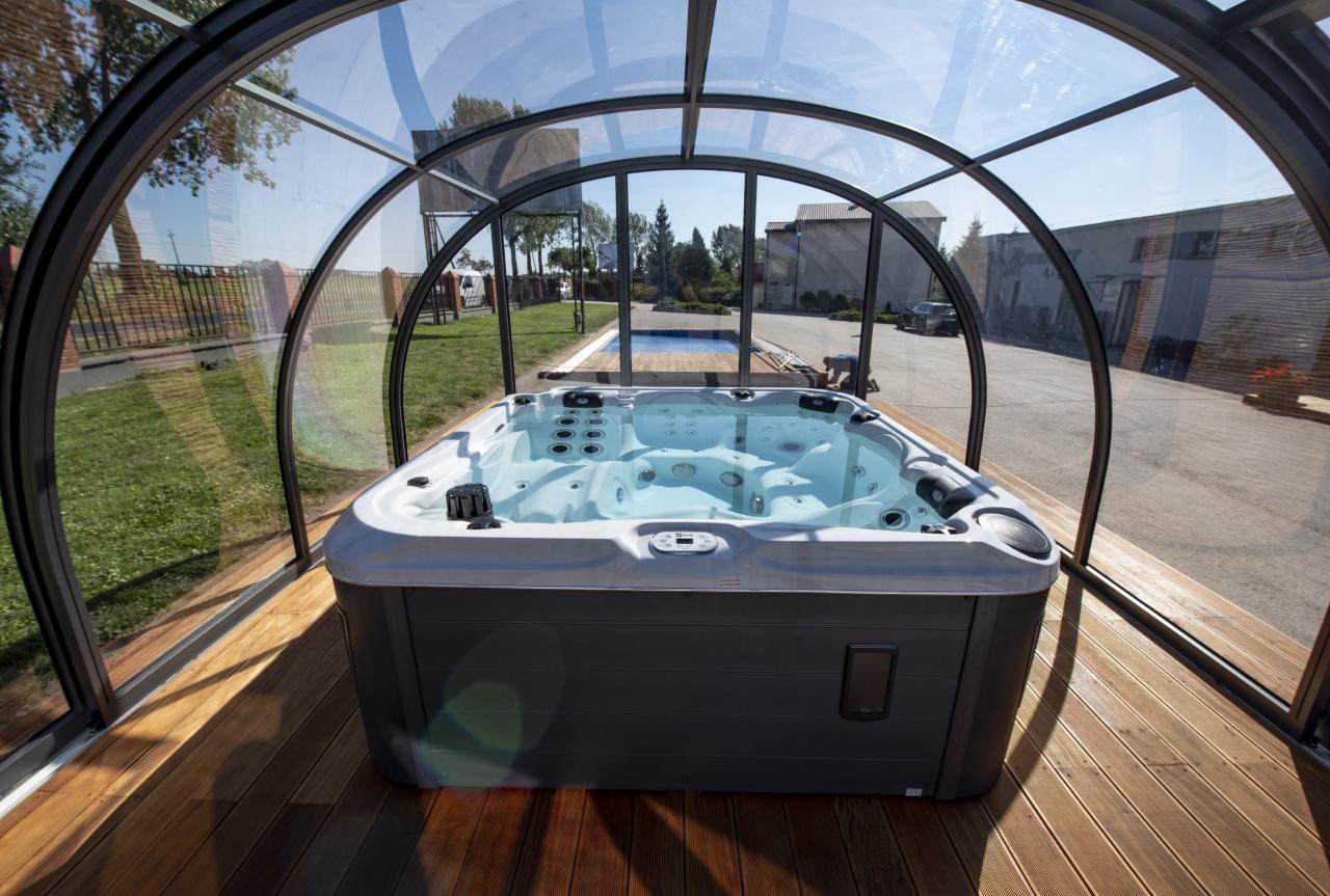 Zamknięta szklaną konstrukcją jacuzzi z hydromasażem, umieszczona na drewnianym tarasie, z widokiem na basen w tle.