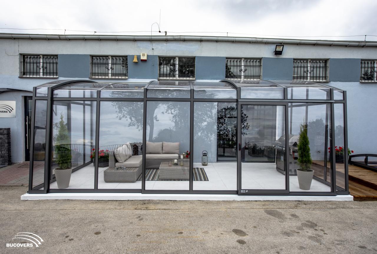 Zadaszony taras z aluminiową konstrukcją i przeszklonymi ścianami, wyposażony w szare meble wypoczynkowe, stolik kawowy, rośliny w donicach, grill i dekoracje, widok z zewnątrz.