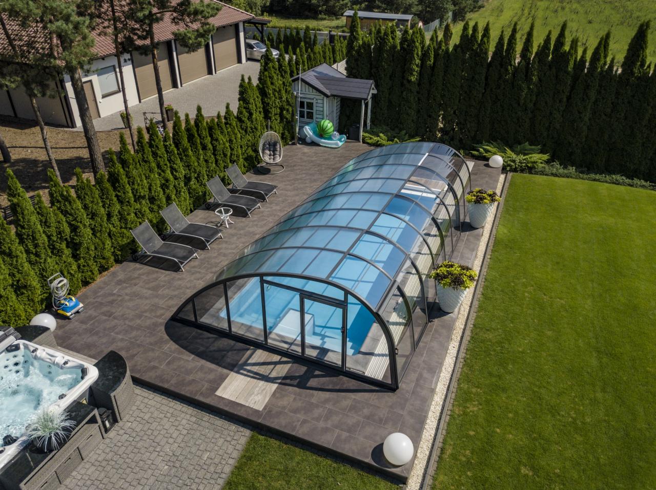 Zadaszony basen z przesuwną konstrukcją na posesji z zielonym trawnikiem, widok z góry, obok jacuzzi, leżaki i altanka.