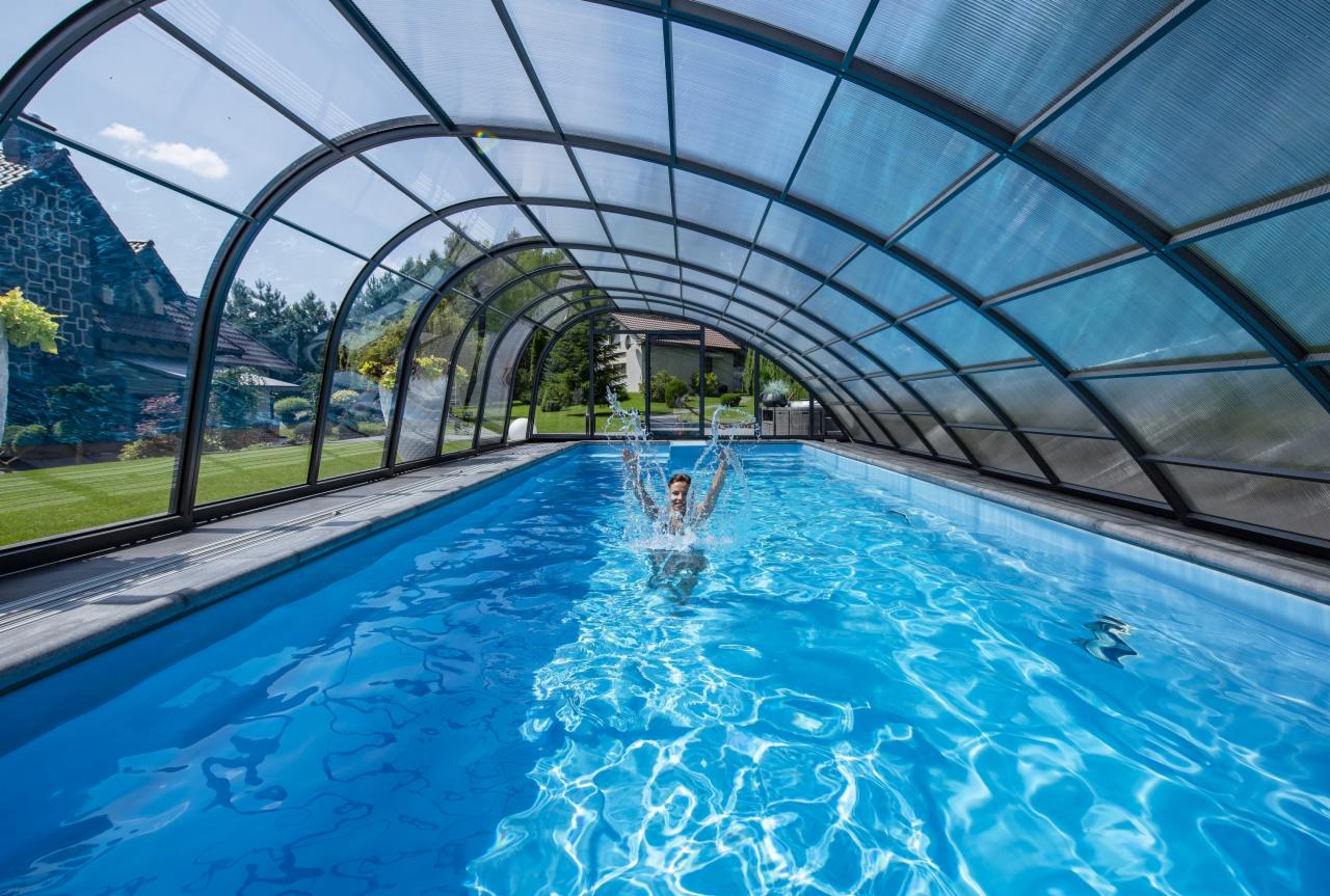 Osoba pluska się w krytym basenie z przezroczystym łukowym zadaszeniem, w tle widoczny dom i zadbany trawnik.