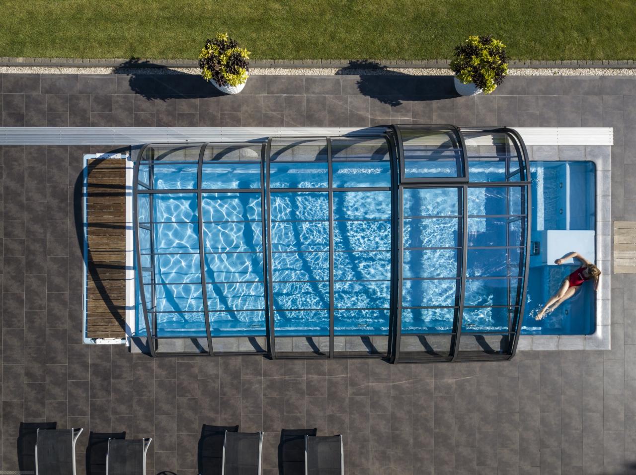 Widok z góry na zadaszony basen z pływającą osobą w czerwonym stroju, otoczony szarą kostką, z dwoma donicami z roślinami i fragmentem trawnika.