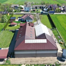 Widok z lotu ptaka na halę z brązowym dachem i szarą elewacją, przylegającą do białej hali namiotowej, na tle wiejskiego krajobrazu z polami i zabudowaniami.