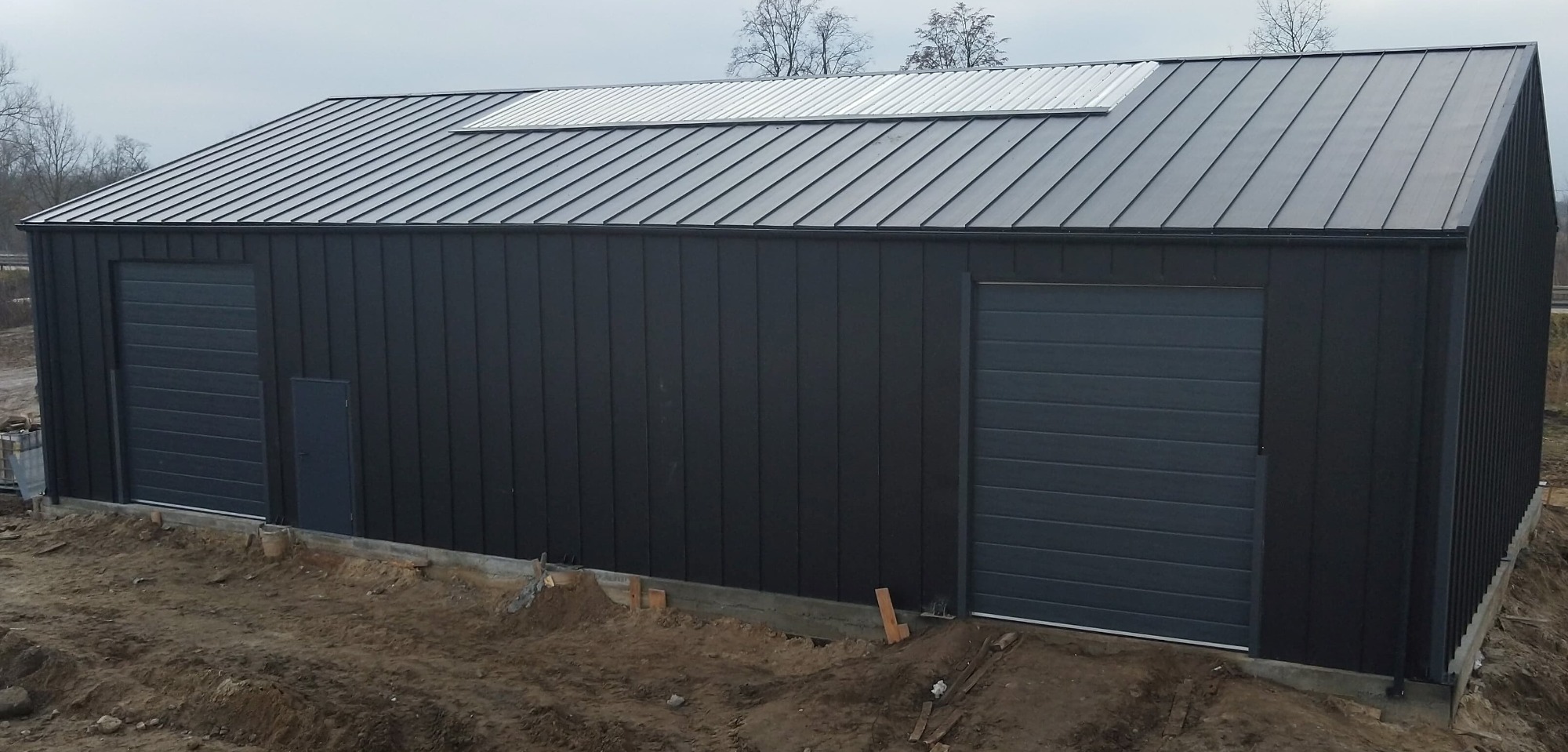 Nowoczesna, ciemnoszara hala z dwoma bramami segmentowymi i panelem świetlnym na dachu, widoczna z zewnątrz na tle ziemi i drzew.