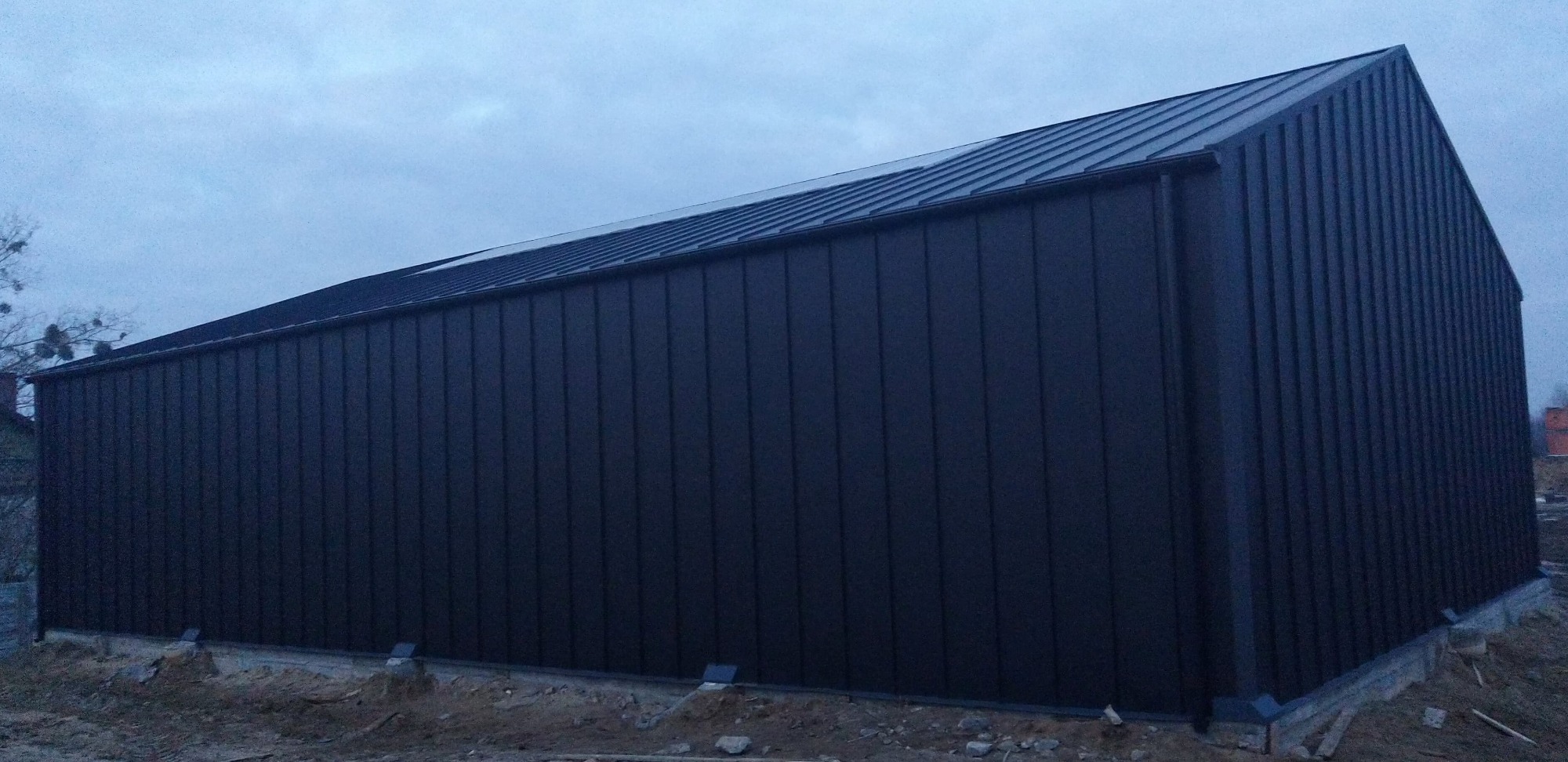 Czarna hala stalowa z blachy trapezowej, widok z zewnątrz przy pochmurnej pogodzie, widać fundamenty i częściowo zagospodarowany teren wokół.