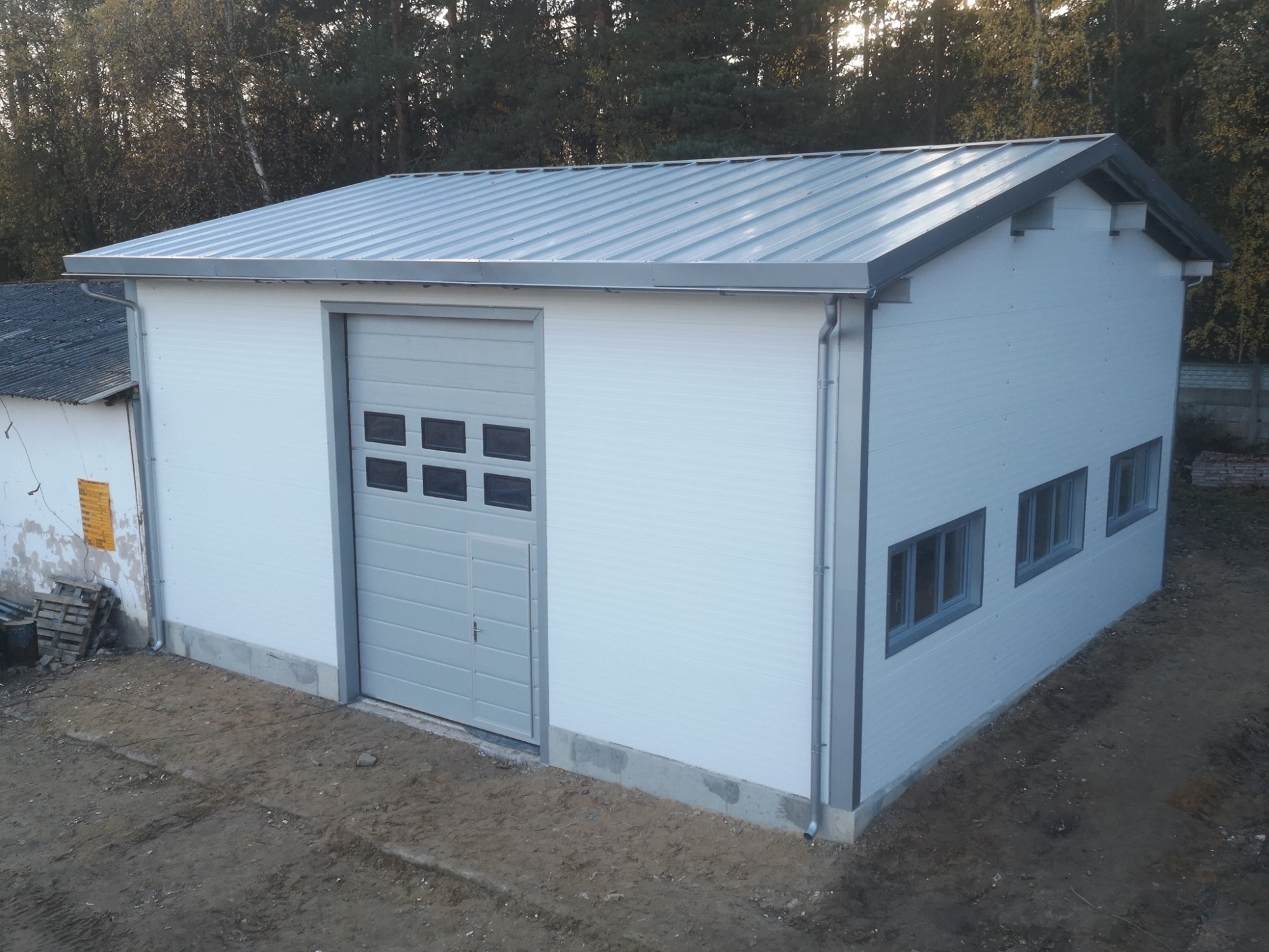 Gotowa hala magazynowa z blachy trapezowej, widoczne okna i brama segmentowa, teren wokół uporządkowany.