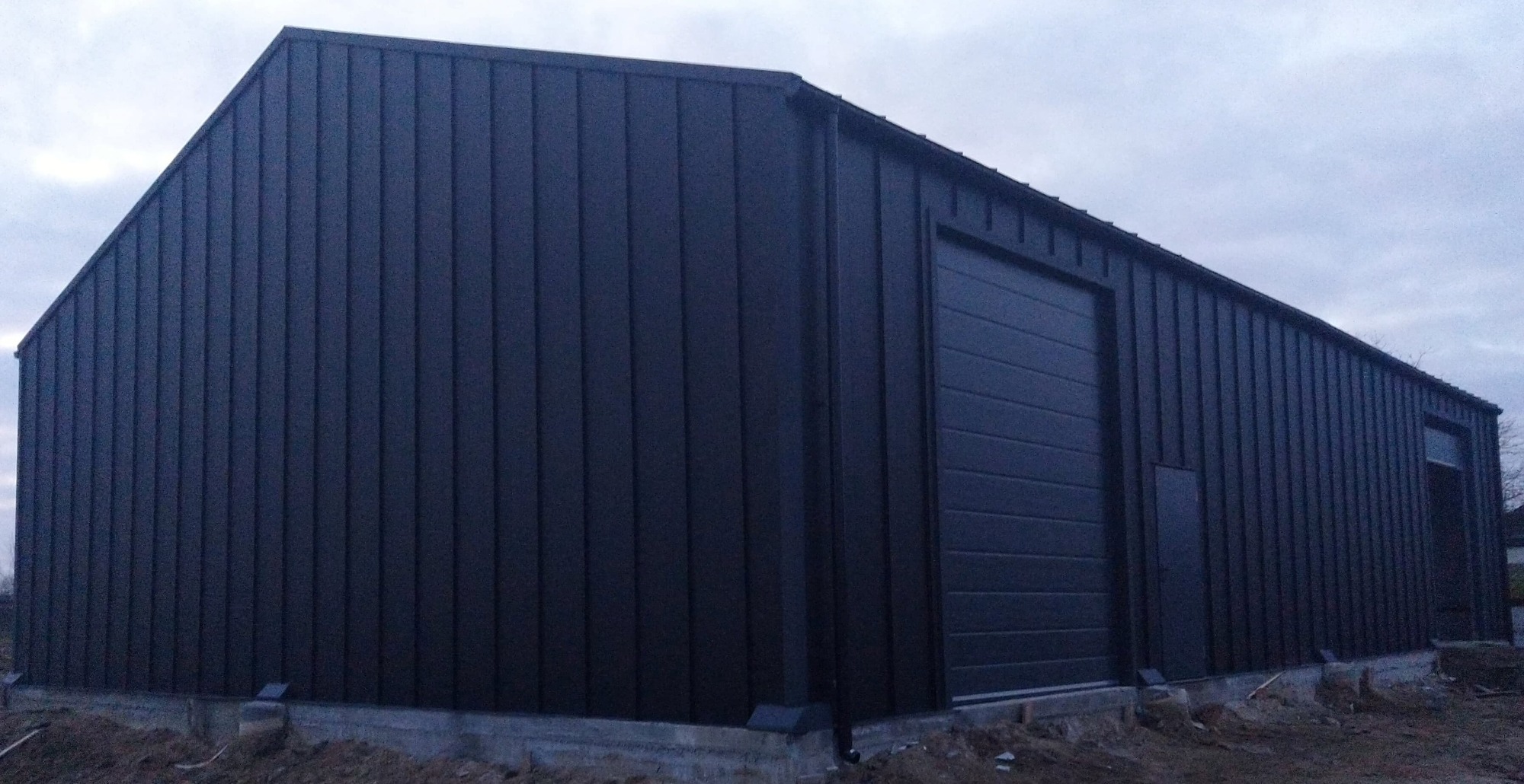 Gotowa hala stalowa z bramą segmentową i pojedynczym oknem, obłożona ciemną blachą trapezową, widoczna podstawa betonowa, pochmurne niebo w tle.