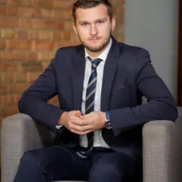 Mężczyzna w ciemnym garniturze i krawacie siedzi w szarym fotelu na tle ściany z czerwonej cegły, dłonie splecione przed sobą, widoczny zegarek na nadgarstku.