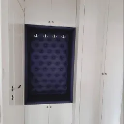 Biała szafa wnękowa z siedziskiem tapicerowanym granatową tkaniną w romby i wieszakami na ubrania, uzupełniona szafkami górnymi i dolnymi.