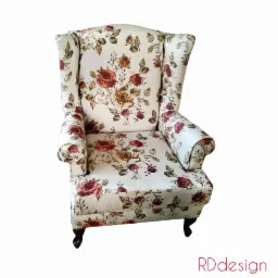 Kremowy fotel typu uszak obity tkaniną w motywy róż i liści na białym tle, widoczne ciemne, toczone nóżki oraz logo RDdesign w prawym dolnym rogu.