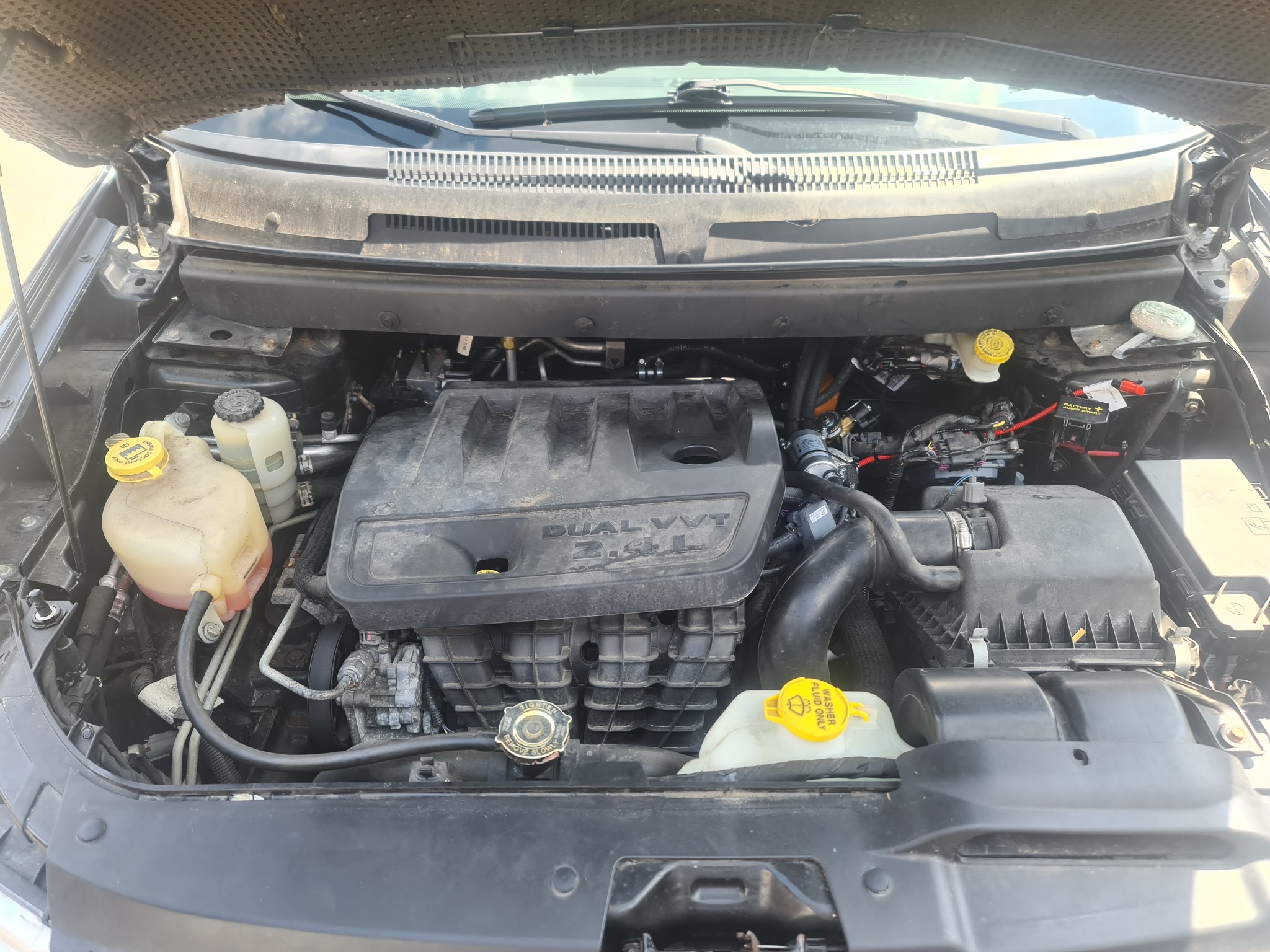 Otwarta maska samochodu z widocznym silnikiem Dual VVT 2.4L, zbiornik płynu do spryskiwaczy z żółtą nakrętką, elementy instalacji. Widoczne ślady użytkowania.