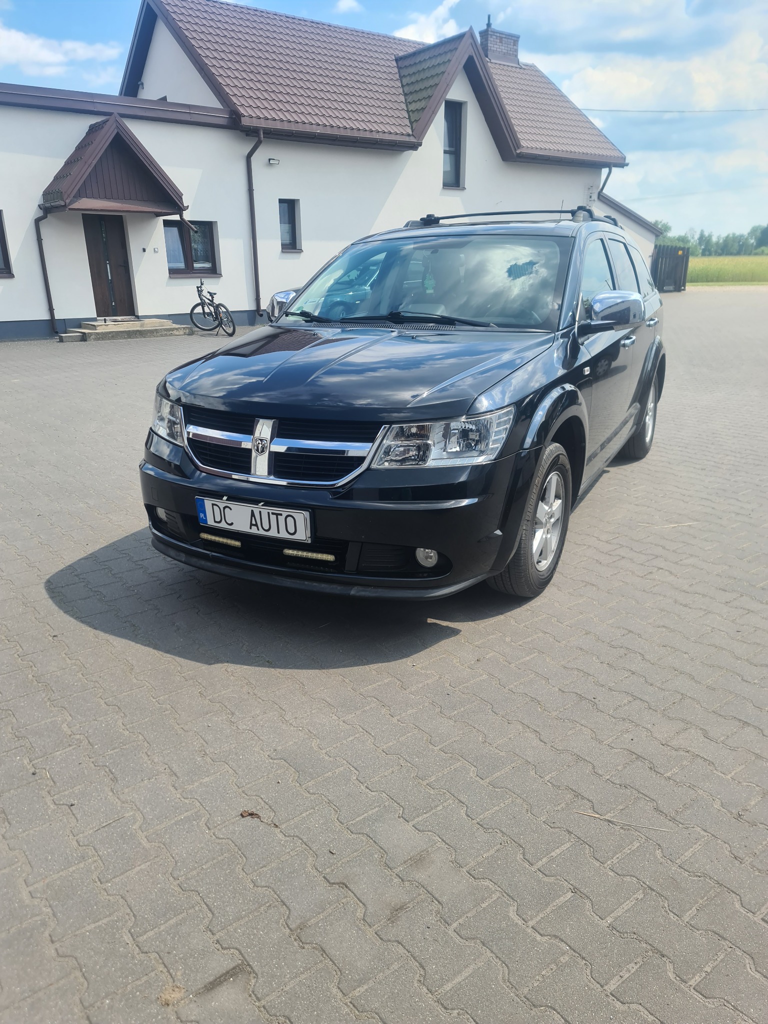 Czarny Dodge Journey zaparkowany na kostce brukowej przed białym domem z brązowym dachem, widoczny rower oparty o ścianę budynku.
