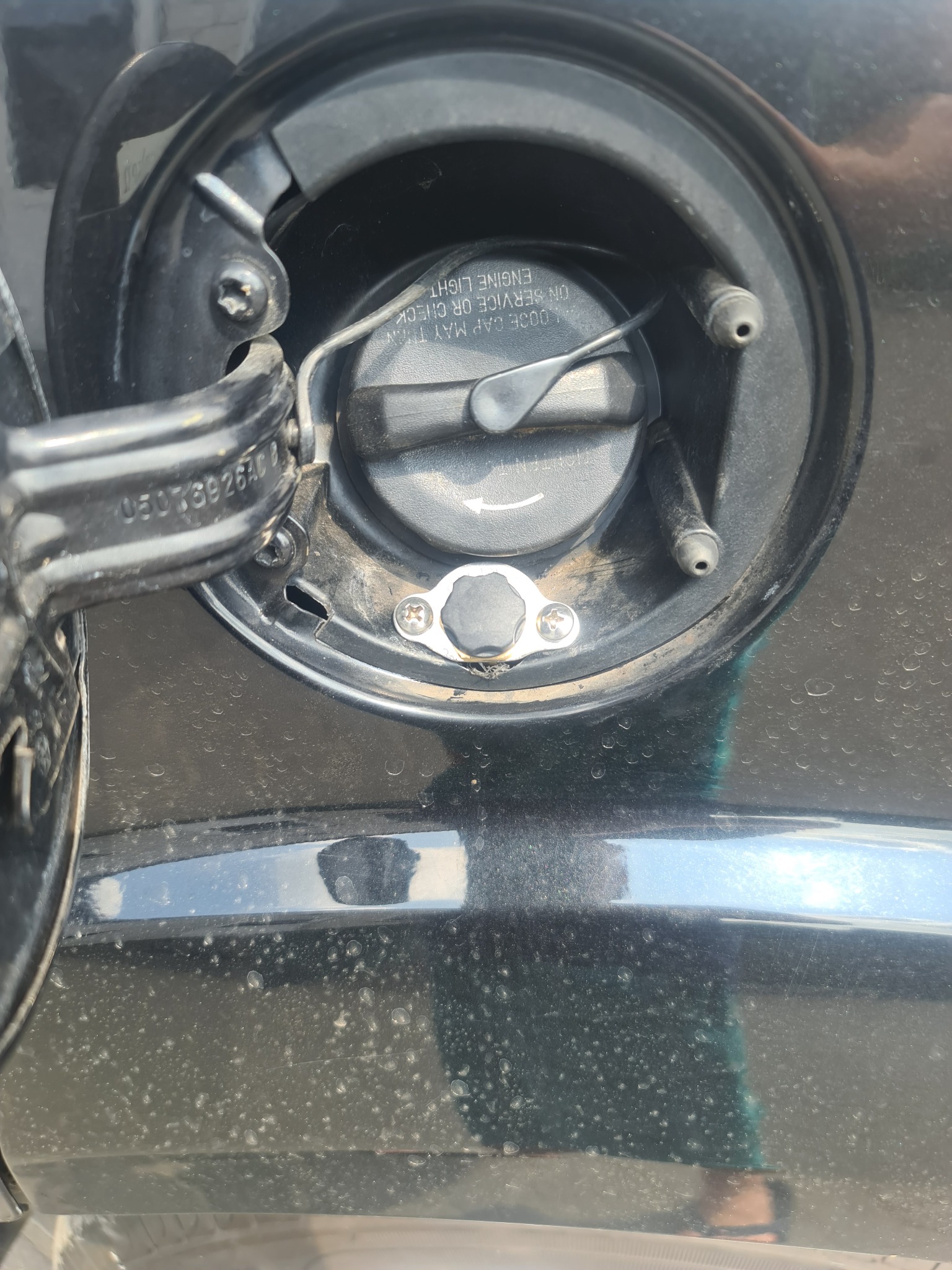 Zbliżenie na wlew paliwa/gazu w samochodzie, widoczne ślady zabrudzeń na karoserii i czarny korek z instrukcją.