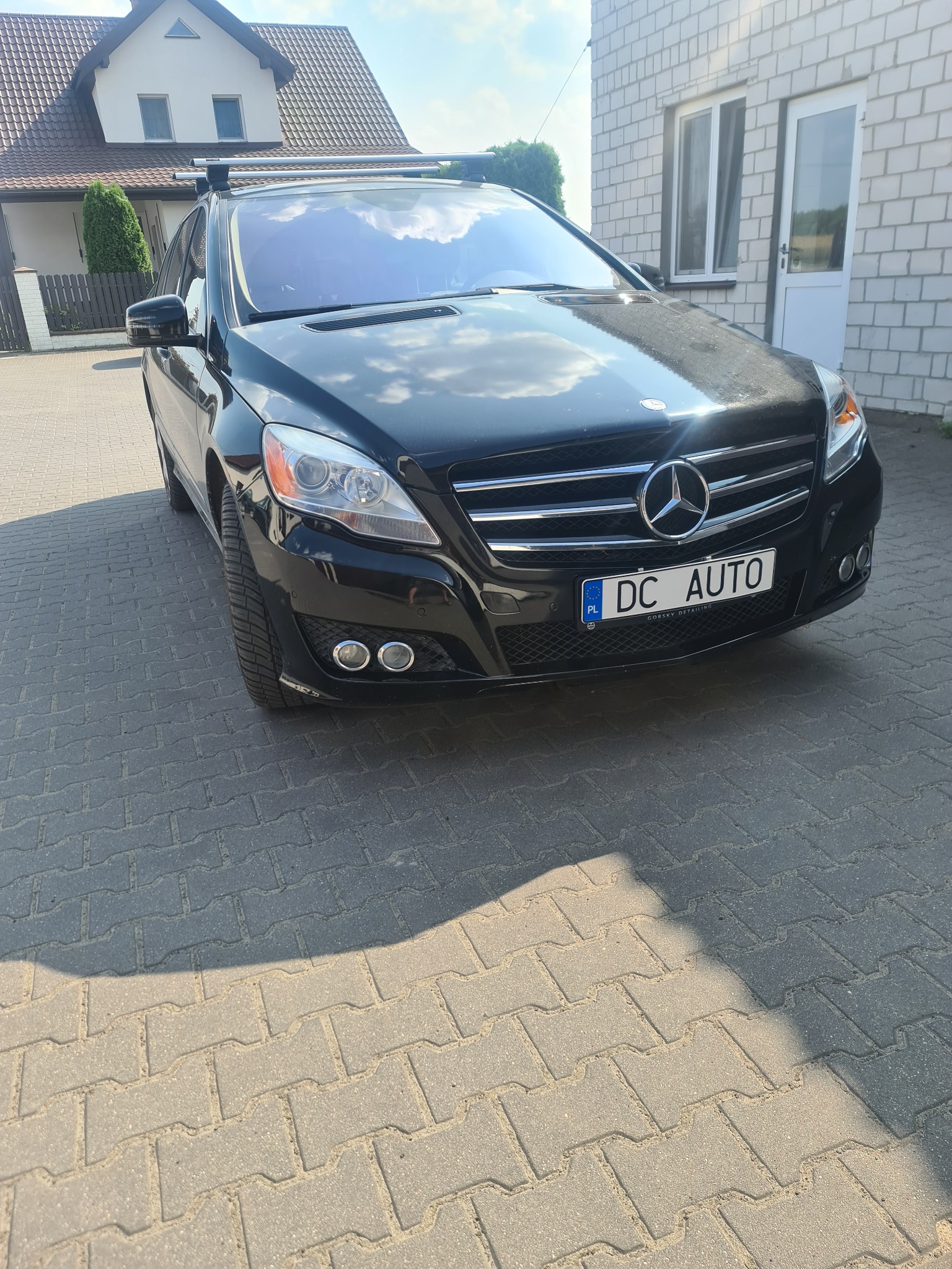 Czarny Mercedes R-klasy z relingami dachowymi zaparkowany na kostce brukowej przed budynkiem, widoczna tablica rejestracyjna z nazwą 'DC AUTO'.