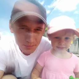 Uśmiechnięty mężczyzna w czapce z małą dziewczynką w różowej sukience i czapce, obok czarnego ogrodzenia, na tle błękitnego nieba.