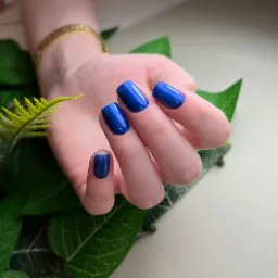 Dłoń z krótkimi, kwadratowymi paznokciami pomalowanymi na intensywny kobaltowy kolor, ozdobiona złotą bransoletką, ułożona na tle z zielonych liści.