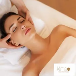 Kobieta z zamkniętymi oczami leży na białym ręczniku podczas masażu twarzy, wykonywanego przez masażystę; widoczne logo salonu kosmetycznego w prawym dolnym rogu.