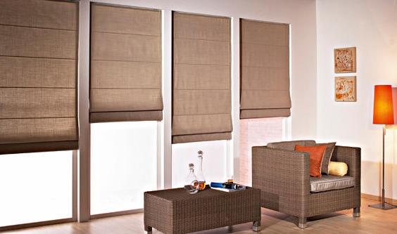 Eleganckie, brązowe rolety rzymskie w trzech oknach, uzupełnione rattanowym stolikiem z dekoracjami i dopasowanym fotelem z poduszkami w salonie z oświetleniem.