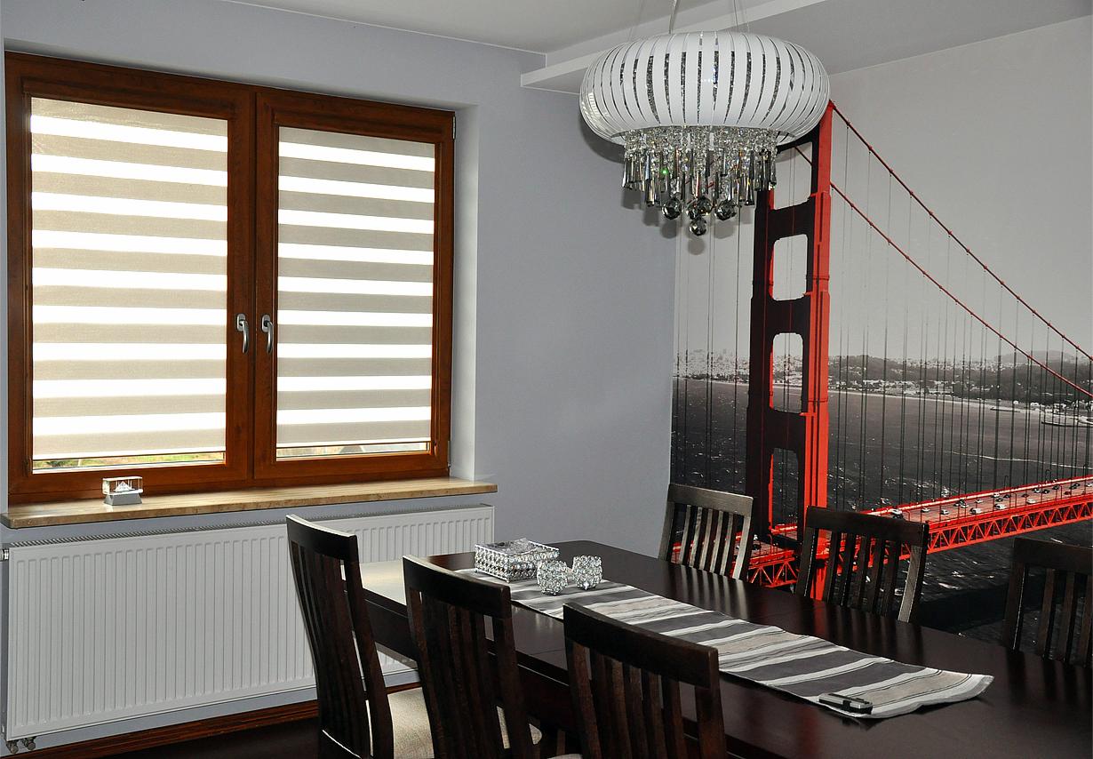 Jadalnia z oknem wyposażonym w rolety w brązowej ramie, na parapecie ozdobny drobiazg, na ścianie fototapeta z mostem Golden Gate, nad stołem designerski żyrandol.
