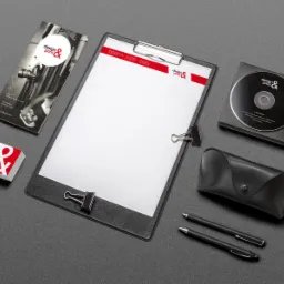 Zestaw materiałów firmowych na szarym tle: wizytówki, ulotka, notes z logo, płyta CD w opakowaniu, etui na okulary i dwa długopisy.