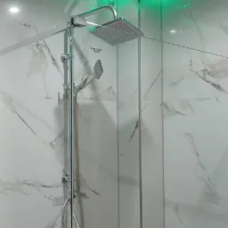 Nowoczesna kabina prysznicowa z deszczownicą, szklanymi ścianami i białymi płytkami imitującymi marmur, podświetlona zielonymi diodami LED na suficie.