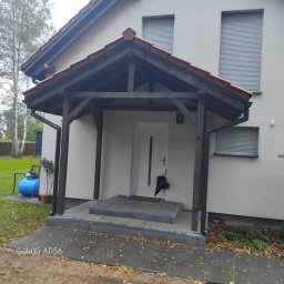 TRANSBUD BLOCH SPÓŁKA Z OGRANICZONĄ ODPOWIEDZIALNOŚCIĄ - Zadaszone wejście do domu z drewnianą konstrukcją i schodami z szarej kostki. Widoczne okna z roletami, trawnik i drzewa w tle. Jesienne liście na chodniku.