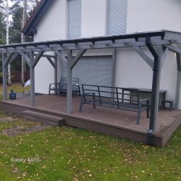 TRANSBUD BLOCH SPÓŁKA Z OGRANICZONĄ ODPOWIEDZIALNOŚCIĄ - Szara drewniana pergola z zadaszeniem i meblami ogrodowymi na brązowym tarasie kompozytowym, na tle białego domu z szarymi roletami i zielonego trawnika z liśćmi.