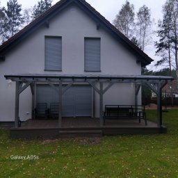 TRANSBUD BLOCH SPÓŁKA Z OGRANICZONĄ ODPOWIEDZIALNOŚCIĄ - Szary dom z zadaszonym tarasem i meblami ogrodowymi. Widoczne rolety w oknach i opadłe liście na trawniku. Ujęcie z poziomu trawy.