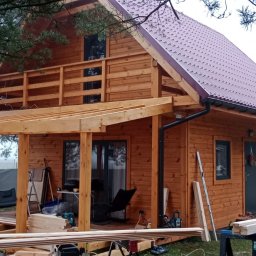 TRANSBUD BLOCH SPÓŁKA Z OGRANICZONĄ ODPOWIEDZIALNOŚCIĄ - Drewniany dom z czerwonym dachem w trakcie budowy, widoczne elementy konstrukcyjne werandy i narzędzia. Tło stanowią drzewa i mglista pogoda.