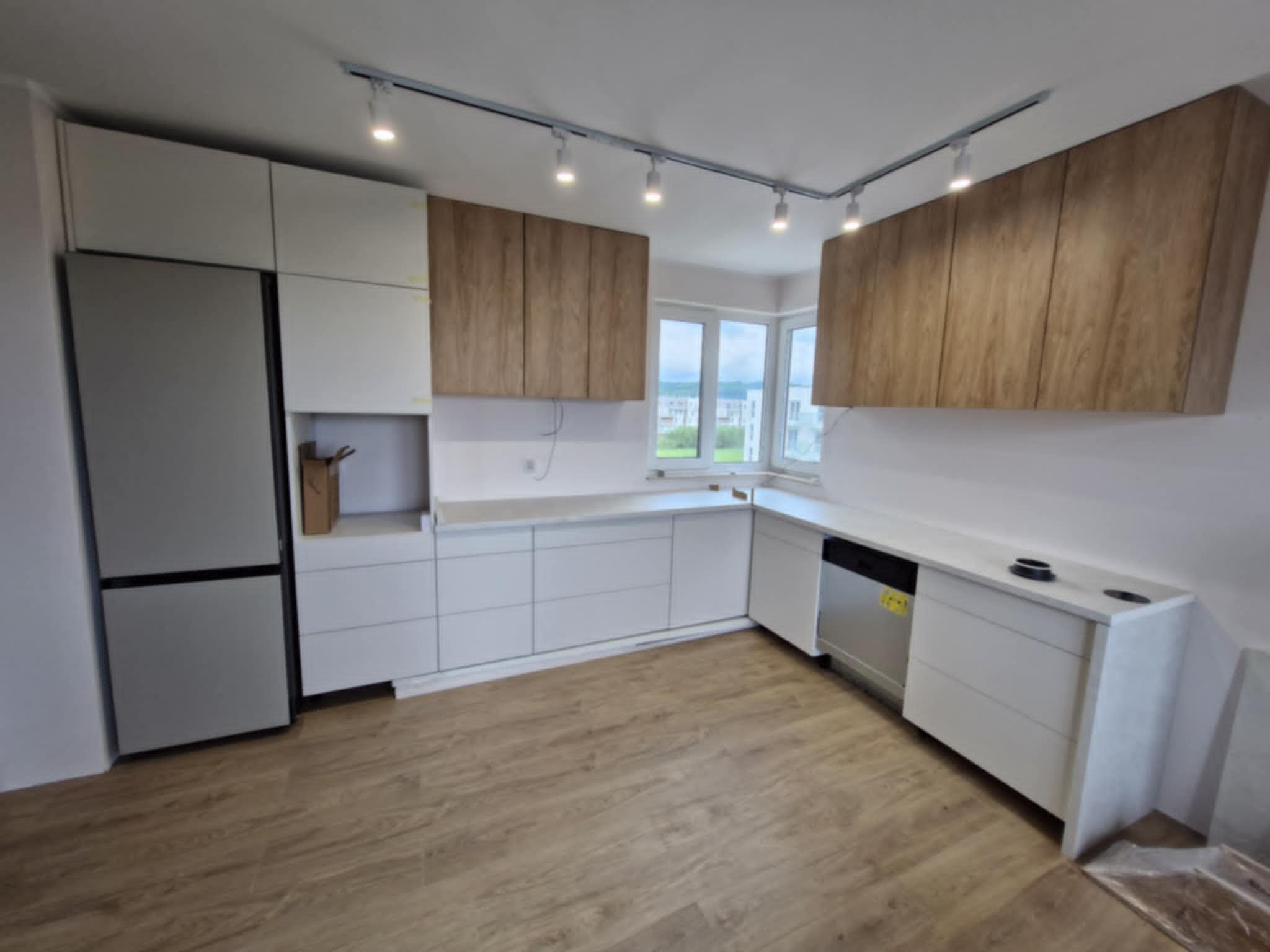 Nowoczesna kuchnia z białymi szafkami dolnymi i drewnopodobnymi górnymi, oświetlona szynoprzewodem. Widoczne okno i podłoga imitująca drewno.