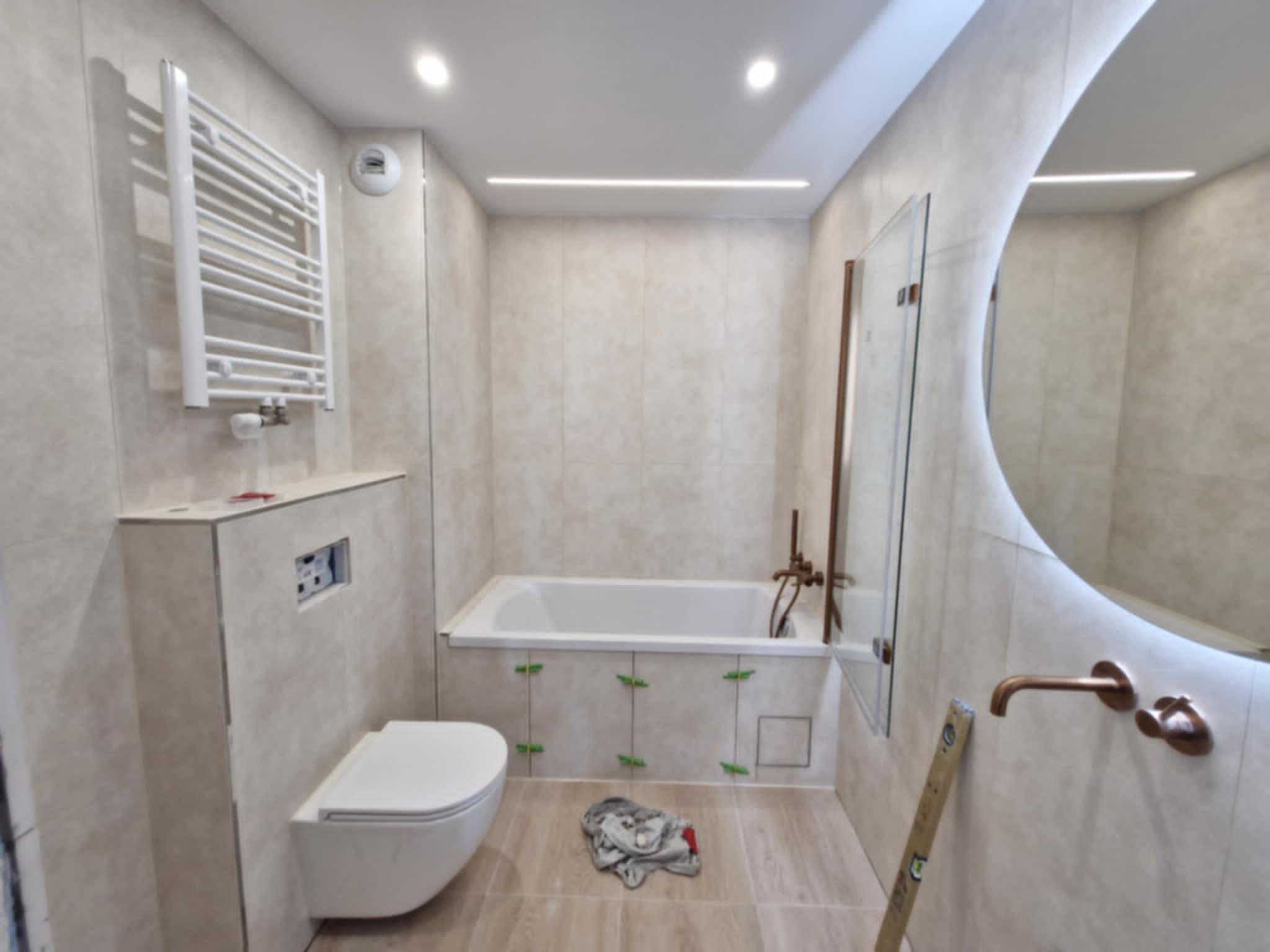 Jasna łazienka w trakcie wykończenia: wanna z zielonymi krzyżykami dystansowymi, toaleta podtynkowa, okrągłe lustro z podświetleniem LED, poziomica.