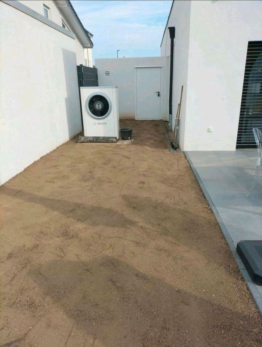 Przygotowany teren pod montaż pompy ciepła Bosch, widok z boku. Białe ściany budynku w tle, teren wyrównany piaskiem.