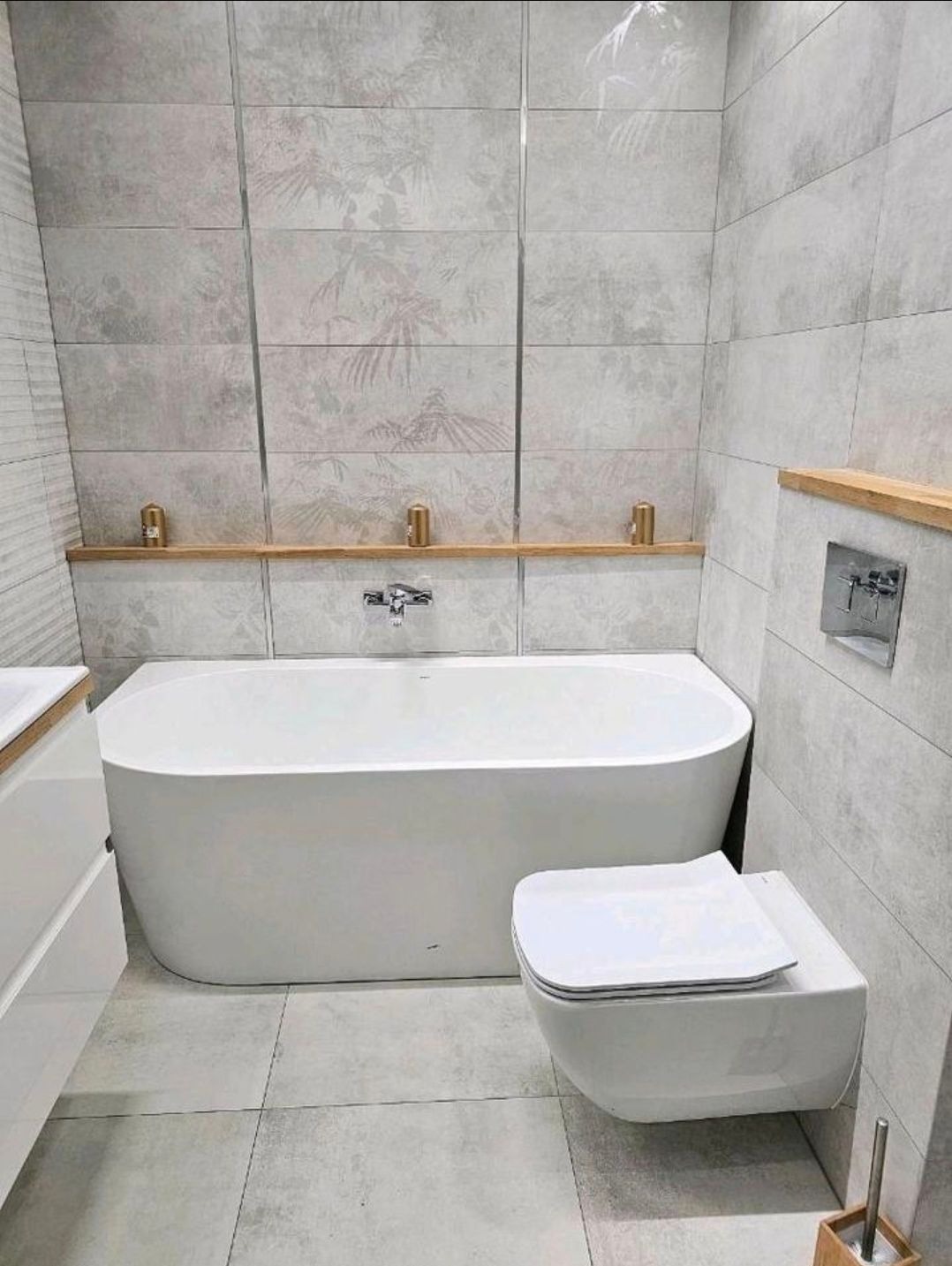Nowoczesna łazienka z wanną, podwieszaną toaletą i szarymi płytkami z motywem roślinnym. Drewniane akcenty dodają ciepła minimalistycznemu wnętrzu.