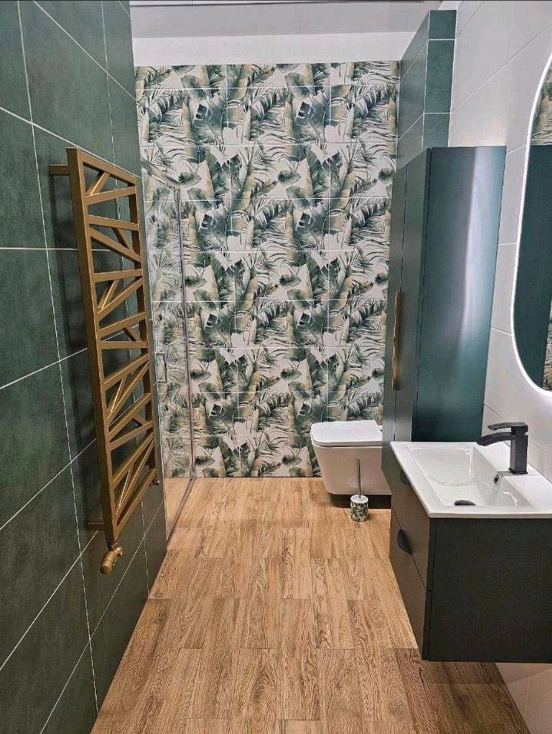 Nowoczesna łazienka z zielonymi płytkami, drewnopodobną podłogą i ścianą w tropikalne liście. Złoty grzejnik drabinkowy i ciemna szafka pod umywalką.