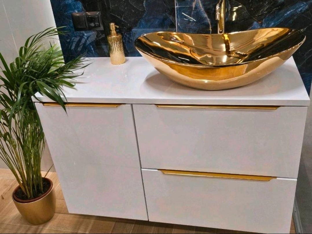Biała szafka łazienkowa z złotą umywalką w kształcie łodzi i złotymi uchwytami, uzupełniona o złotą doniczkę z rośliną i dozownik na tle marmurowej ściany.