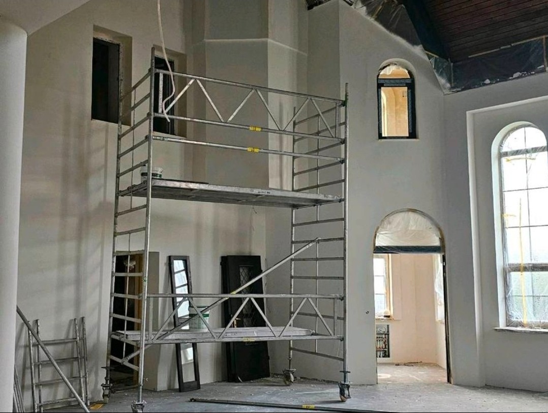 Wnętrze w trakcie prac wykończeniowych: rusztowanie na tle świeżo wyszpachlowanych ścian z oknami o łukowatych ramach. Widoczne przygotowania do malowania.