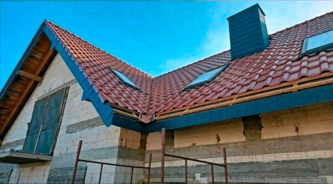 Dom w trakcie budowy z czerwoną dachówką, niebieskimi elementami wykończeniowymi i oknami dachowymi. Widoczne rusztowania i niezakończona elewacja.