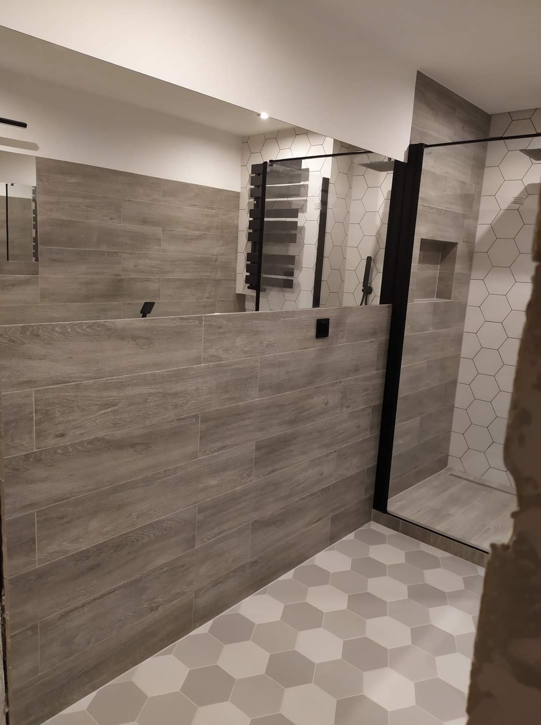 Wykończona łazienka z drewnopodobnymi płytkami na ścianach, heksagonalną podłogą i czarną kabiną prysznicową. Widoczny grzejnik drabinkowy i wnęka w ścianie.