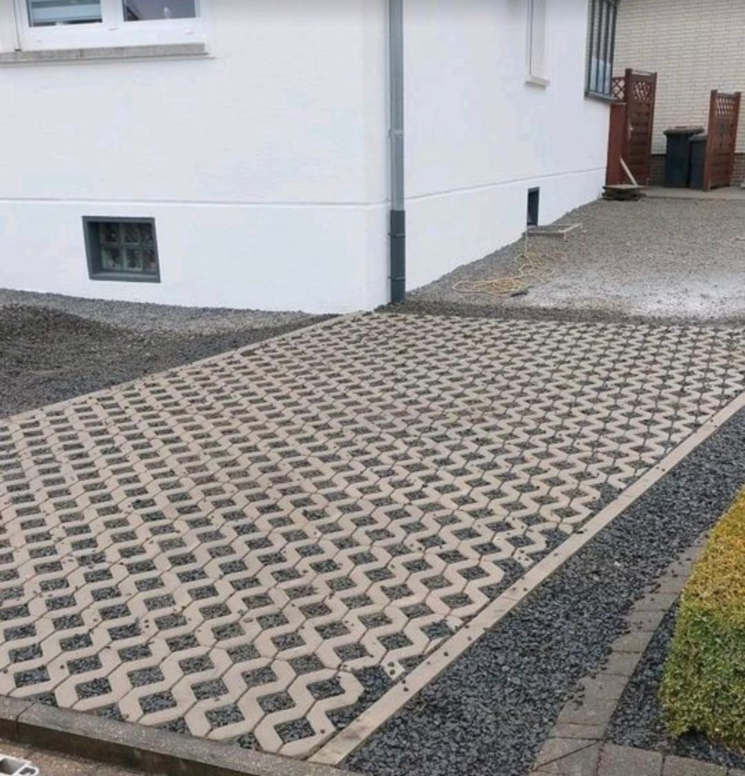 Ułożona ażurowa kostka brukowa na podjeździe, z wypełnieniem żwirowym, obok białego budynku z szarą rynną. Widok z góry, lekki kąt.