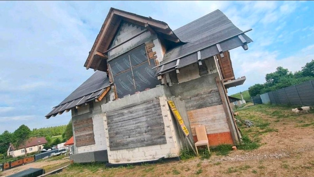 Niedokończony dom z drewnianym szalunkiem okiennym i czarną membraną na poddaszu, widok z boku na tle zieleni i błękitnego nieba.