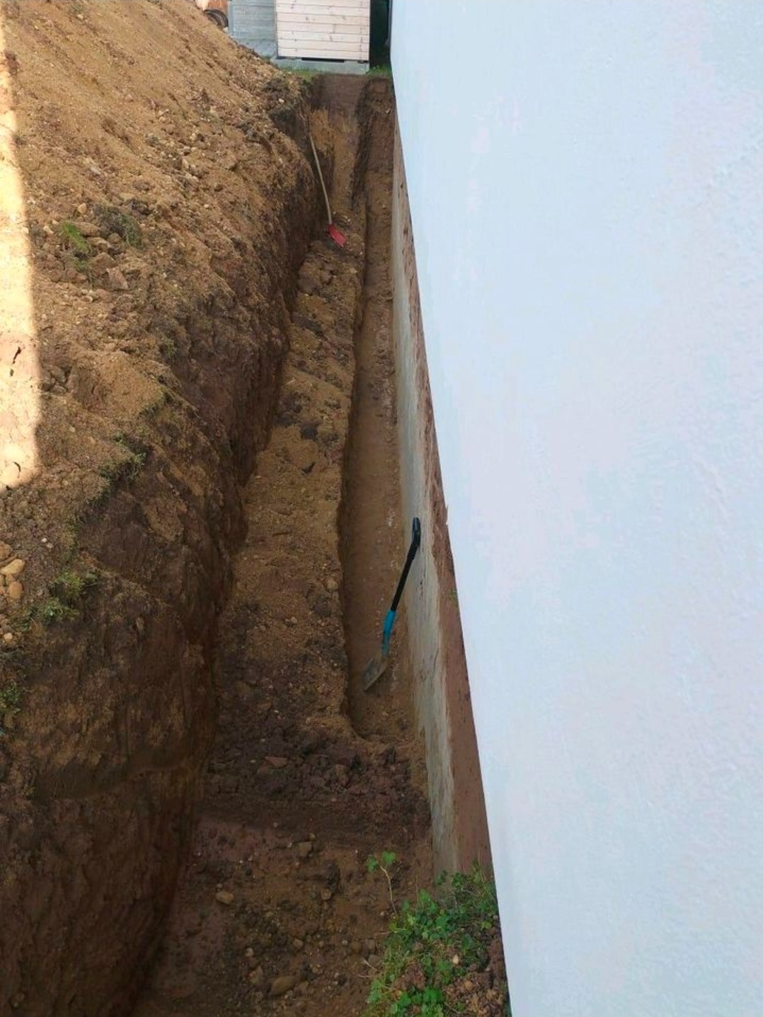 Wykop wzdłuż ściany budynku, widoczne narzędzia (łopata, szpadel). Przygotowanie do prac ziemnych, prawdopodobnie związanych z izolacją fundamentów.