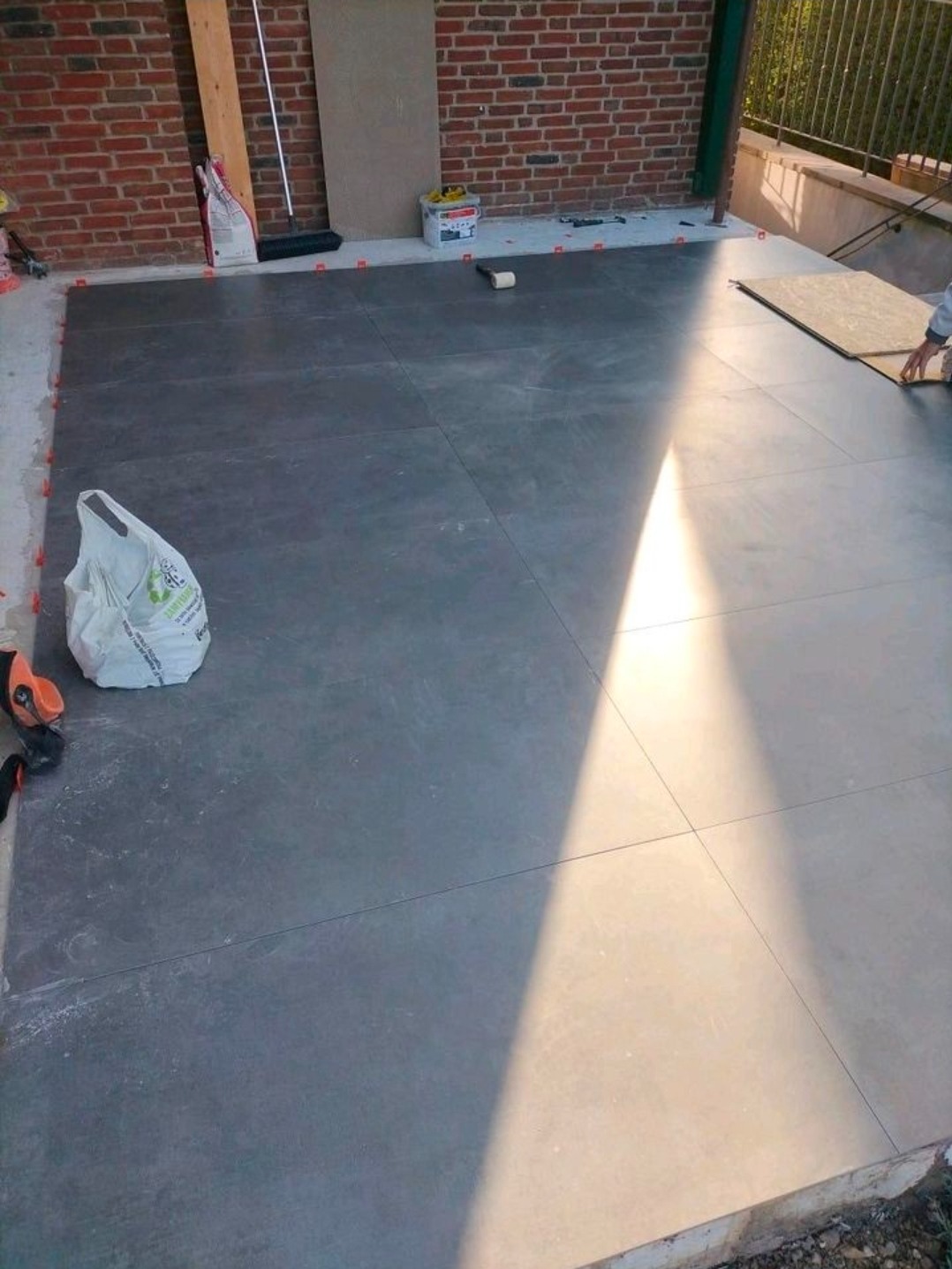 Układanie ciemnych, kwadratowych płytek na tarasie. Widoczne narzędzia, poziomice i krzyżyki dystansowe. Częściowo ułożona podłoga, ceglana ściana w tle.