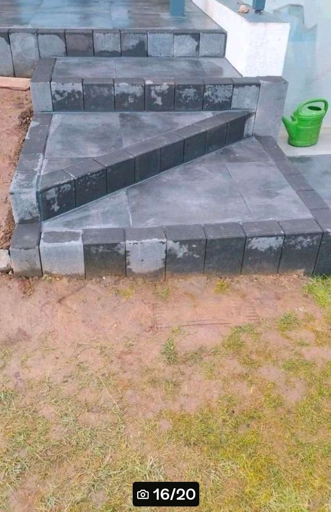 Schody wejściowe wyłożone szarą kostką brukową z ciemnymi obrzeżami, z zieloną konewką w tle, na zewnątrz budynku. Widok z góry na fragment trawnika.