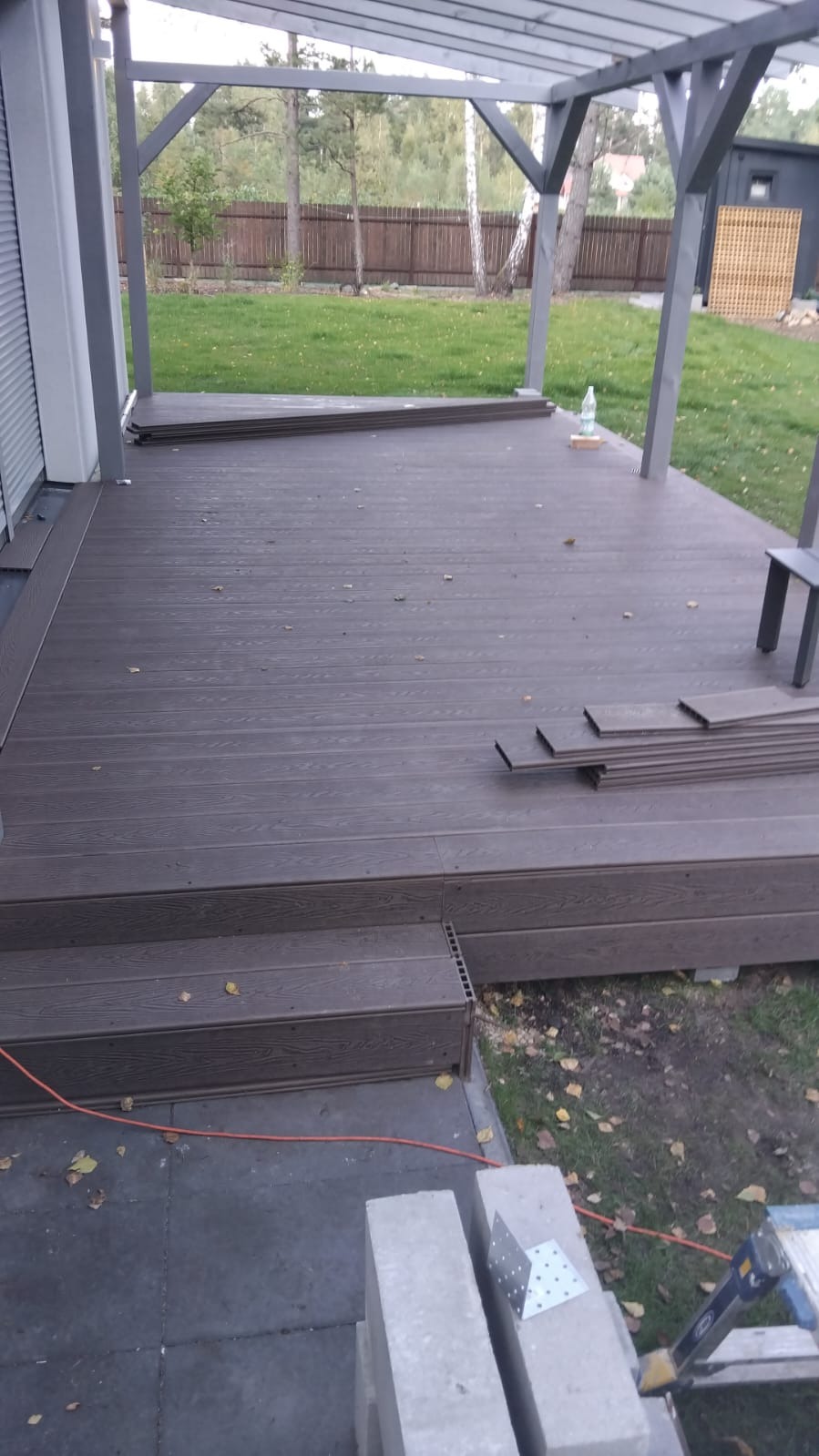Gotowy taras z deski kompozytowej w odcieniach brązu, z widocznymi schodkami i elementami konstrukcji zadaszenia. Na tarasie leżą dodatkowe deski.