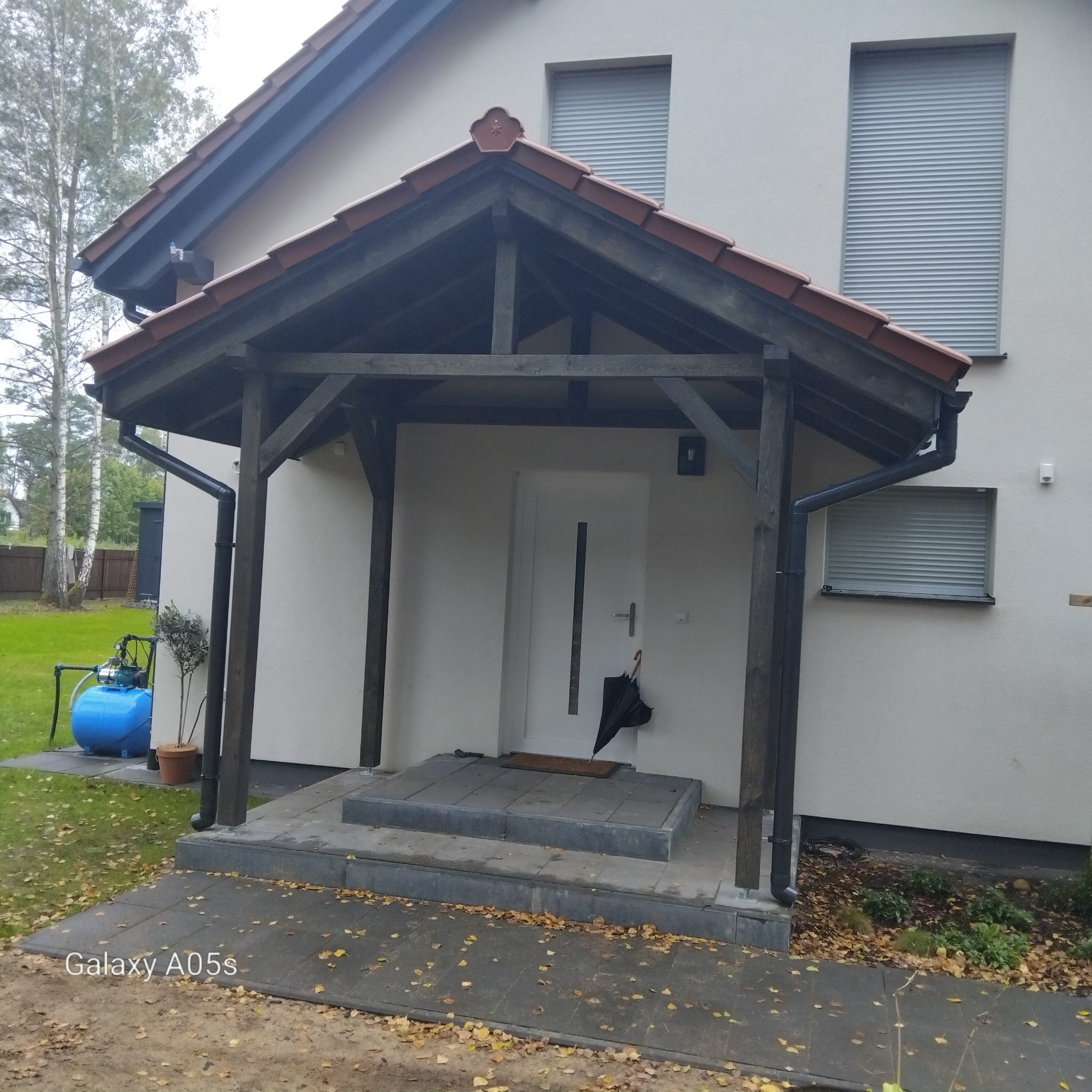 Zadaszone wejście do domu z drewnianą konstrukcją i schodami z szarej kostki. Widoczne okna z roletami, trawnik i drzewa w tle. Jesienne liście na chodniku.